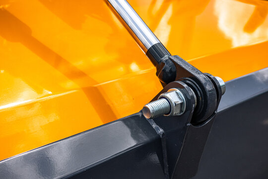 Hydraulic cylinder rod and pivot joint on heavy agricultural machinery. Steel piston connection detail with threaded bolt, nut fastener, hinge assembly, and yellow industrial metal frame.