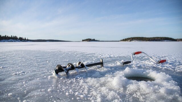 Black manual ice auger lying next to a drilled hole on a frozen lake. Winter outdoor recreation and equipment on a snowy surface. Ice fishing hobby and cold weather adventure concept