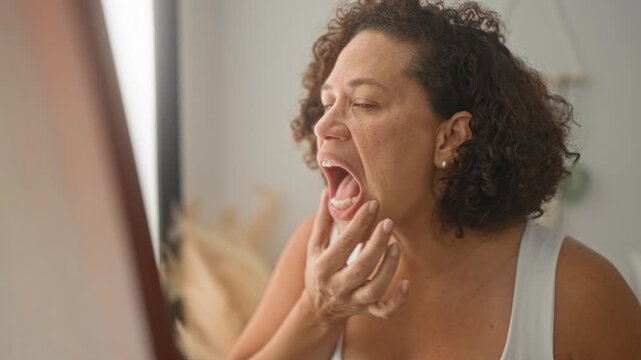 Middle aged woman pinches cheek and inspects mouth and teeth in a studio mirror while touching jaw and opening mouth to check alignment; concern selfcare.