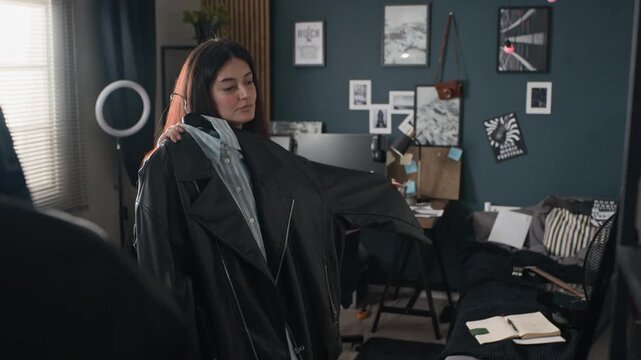 Young adult woman choosing outfit from clothing rack and holding leather jacket in front of mirror. Perfect for fashion retail promotions, lifestyle vlogs, or e-commerce advertising