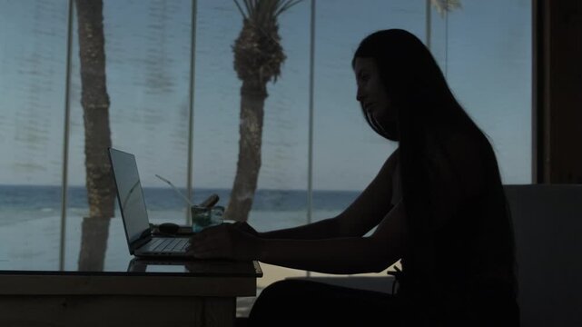 Focused female freelancer working on laptop in tropical cafe with ocean view, concentrated on her work in a relaxed coastal setting, remote work and digital nomad lifestyle