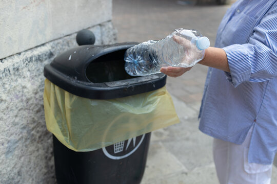 person's hand throwing an empty transparent plastic water bottle into a black public trash bin on a city street. Concept of waste management, recycling plastic, environmental care, anti-pollution, and