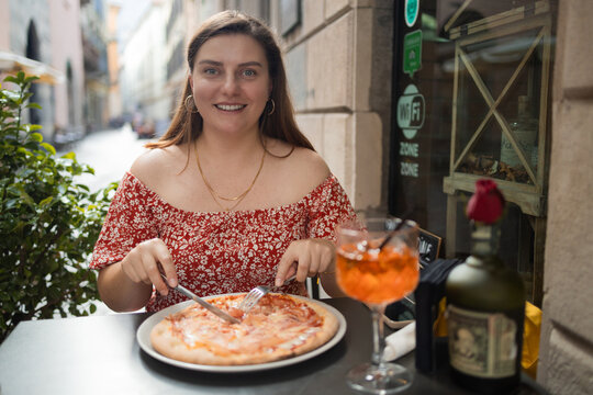 smiling beautiful young woman enjoying a slice of fresh Italian pizza at a cozy outdoor restaurant table. She is having lunch with a traditional orange cocktail on a narrow picturesque European street