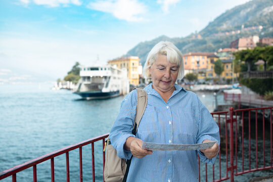 Smiling elderly woman traveler holding a paper map while standing on the waterfront promenade in Varenna, Lake Como, Italy.