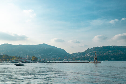 A breathtaking wide shot of Lake Como (Lago di Como) in Lombardy, Italy. The scene showcases vibrant blue water with moving boats, charming coastal town architecture nestled on hills, and green