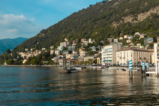 A breathtaking wide shot of Lake Como (Lago di Como) in Lombardy, Italy. The scene showcases vibrant blue water with moving boats, charming coastal town architecture nestled on hills, and green