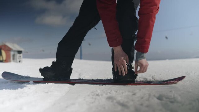 A person secures their bindings on a snowboard, preparing for a winter ride. The individual fastens their boots to the board, ready for snowboarding on the snow.
