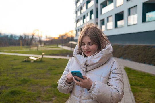 A woman is smiling and looking at the camera in a grassy outdoor area with a modern building in the background. The scene conveys a happy and relaxed mood, with natural green and white hues.