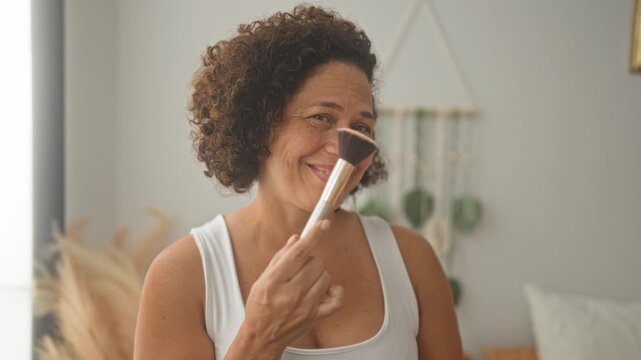 Woman holding makeup brush to cheek in cozy bedroom, smiling and demonstrating skincare routine; self care calm.