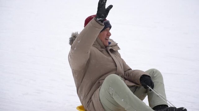 Cheerful senior Asian man sliding down snow hill on a yellow sled at Furano ski resort in Hokkaido, Japan.