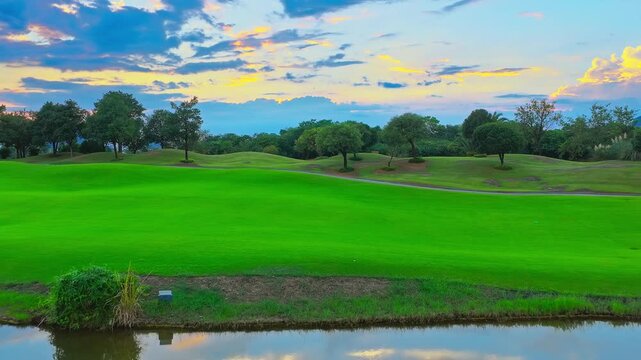 Lush golf course with reflective pond, manicured fairway, mature trees, and colorful sunset clouds, suitable for sports, leisure, tourism, and outdoor backgrounds.
