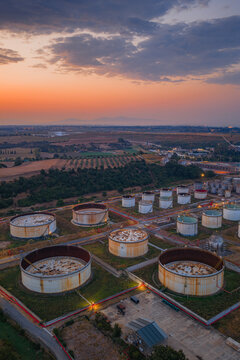 Aerial view of a fuel depot with large cylindrical storage tanks, industrial infrastructure, and surrounding rural landscape under a dramatic sunset sky in Rome, Lazio, Italy.