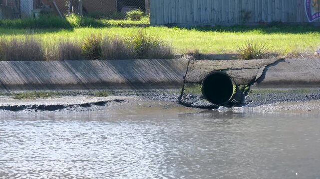a concrete stormwater outfall pipe discharging into the tidal waters of Cherry Creek in Seaholme, Melbourne, Australia.&nbsp;&nbsp;Retaining wall, muddy tidal banks&nbsp;and grassy embankments line the waterway