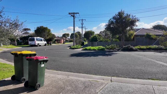 A quiet residential street in Seaholme, Melbourne, Australia, with wheelie bins placed along the kerb for kerbside collection, overhead power utility poles, suburban homes lined the neighborhood road