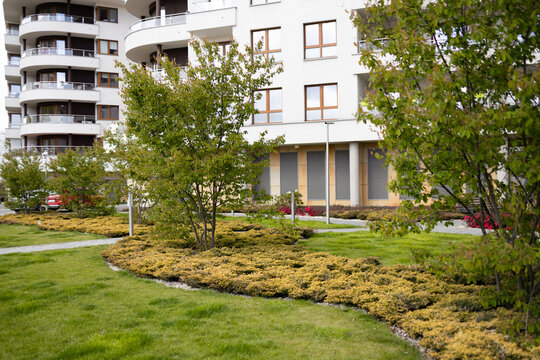 A beautiful scenic view of a modern residential apartment building or alpine style resort hotel. The property features well-manicured green lawns, decorative bushes, and landscaped gardens in the