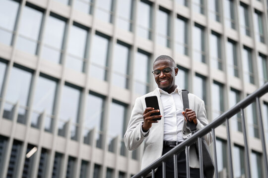 Real estate agent, black businessman, using smartphone, calling, outdoor near office building. African business man smiling, prepared for client meeting, leading cross-functional projects. Copy space
