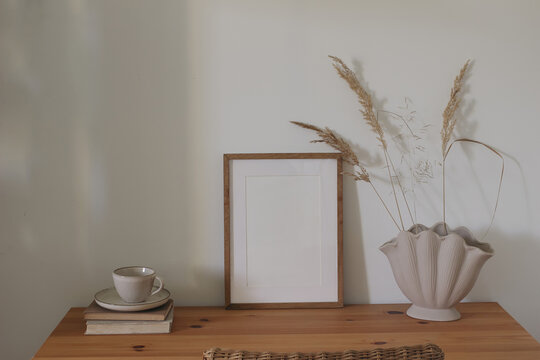 Minimal Scandinavian home interior with blank wooden frame mockup, shell ceramic vase, dried grass, coffee cup and books on wooden table in soft natural sunlight with copy space. Rattan chair.
