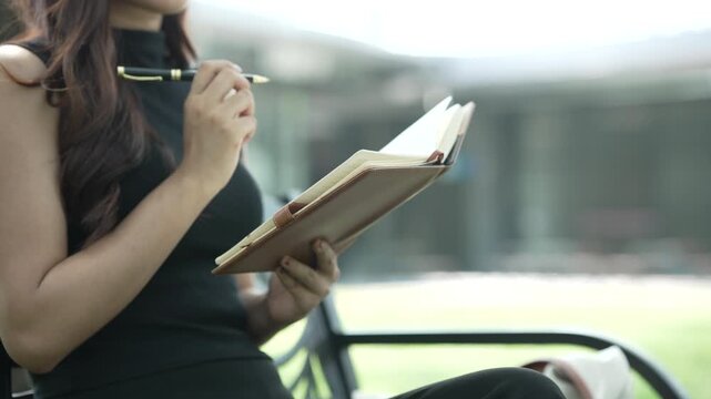 Close view of a young woman in black clothing writing in a notebook while sitting outdoors, with soft light and blurred background, suggesting planning, study, or quiet reflection.