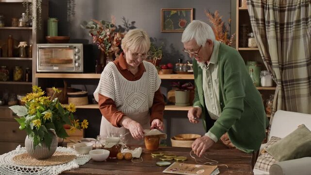 Senior Caucasian woman and man wrapping homemade honey jar with paper and string in rustic kitchen. Video is perfect for retirement lifestyle or organic food brand commercials