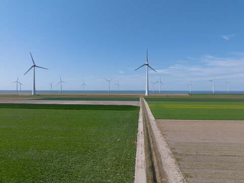 Aerial view of a wind farm with large turbines standing in green agricultural fields and a dirt path leading towards the horizon in the Netherlands.