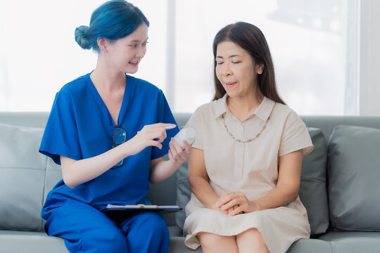 Young Asian female doctor or nurse with blue hair explaining dosage instructions for medication bottle to a senior female patient in a hospital or clinic.