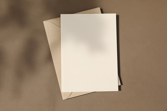 Blank greeting card mockup with kraft envelope on neutral brown table textured background in warm natural sunlight, shadows. Minimalist summer stationery template. Invitation, branding wedding design