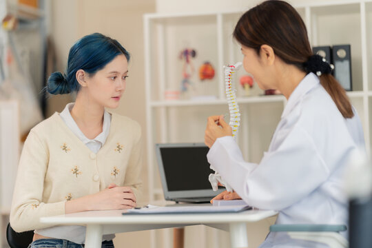 Female doctor explaining spine model to a young patient with blue hair in a clinic, medical consultation about back pain or posture correction.