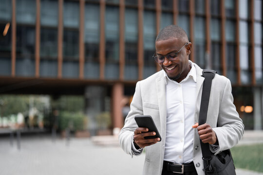 Black businessman working as banking manager, using smartphone, texting, outdoor near office building. African american business man smiling, ready for next meeting, managing accounts. Copy space