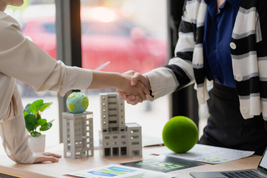 Close up of business partners shaking hands with architectural building models, globe and green concept on office table.