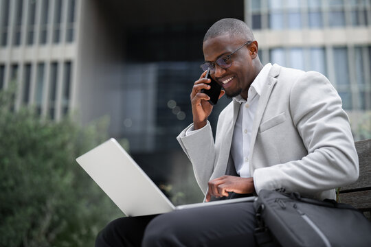 Portrait of african businessman, financial advisor, sitting on bench, using laptop and talking on smartphone, outdoor near office building. Black business man smiling, staying productive. Copy space