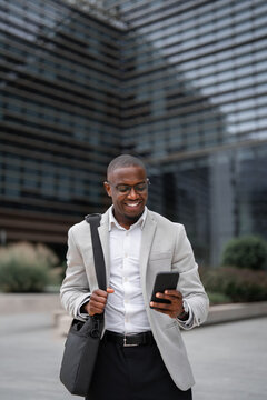 African american businessman working as business consultant, using smartphone, texting, outdoor near office building, vertical. Black business man smiling, staying productive throughout day.