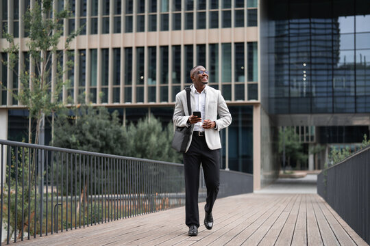 Portrait of black businessman, business owner, walking, using smartphone, checking email, outdoor near office building, fullbody. African business man smiling, staying connected with technology.