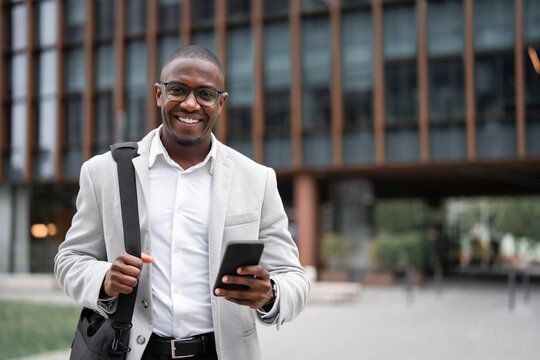 Using smartphone, messaging client, african american businessman outdoor near office building, works as startup founder. Black business man smiling, reviewing project details, managing accounts.