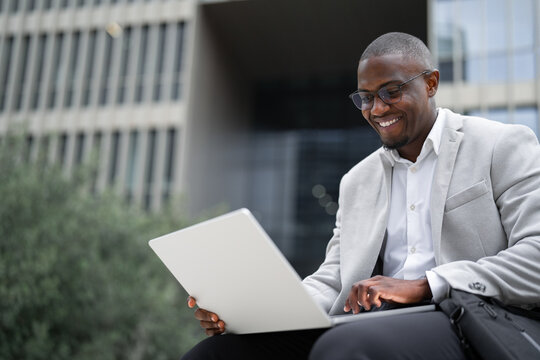 It consultant, african american businessman, sitting, using laptop, typing report, outdoor near office building. Black business man smiling, reviewing project details, managing accounts. Copy space