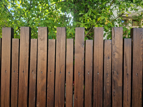 wooden picket fence painted with dark brown protective glaze in a rosewood-like shade with visible wood structure and attached with screws. jagged edge like battlements