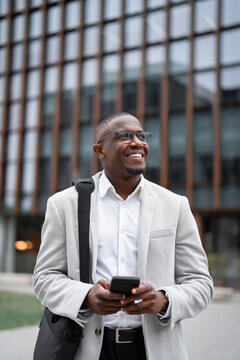 African american businessman working as consultant, using smartphone, texting, outdoor near office building, vertical. Black business man smiling, focused on daily tasks, managing accounts, advancing.