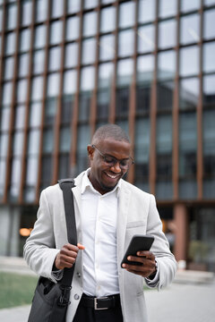 Sales manager, african businessman, using smartphone, calling, outdoor near office building, vertical. Black business man smiling, staying productive throughout day, leading cross-functional projects.
