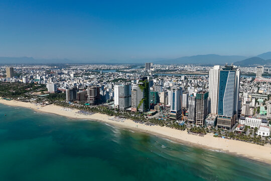 Coastal Da Nang city features many tall buildings next to My Khe Beach, where turquoise ocean water meets golden sand under a clear sky
