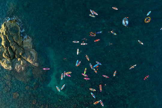 Aerial view shows many people on colorful stand up paddleboards and in kayaks enjoying the clear blue ocean near a large rocky island during daytime