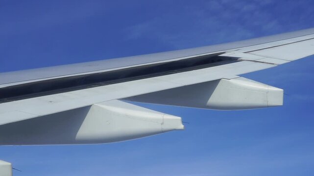 Airplane wing flaps rising and retracting as an airplane travels across a blue sky, as viewed from a window seat.