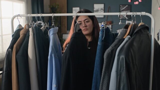 Young adult woman choosing clothes from rack at home, examining blue jeans and shirt. Perfect for fashion retail promotions, wardrobe organization apps, or personal styling videos