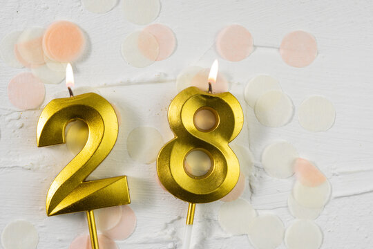 Twenty-eighth anniversary celebration. A golden burning candle in the shape of the number twenty-eight on a festive white background with confetti and glitter. Festive banner.