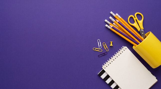 Top view flat lay of school and office stationery including yellow pencils, scissors, notepad, and paperclips on a vibrant purple background with copy space.