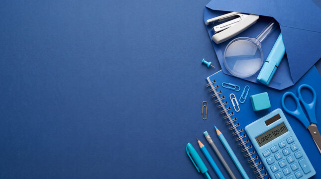 Flat lay top view of blue office supplies including a notebook, calculator, scissors, pens, envelope, and magnifying glass arranged on a blue background with negative space for text.