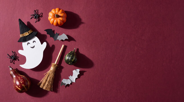 Flat lay Halloween composition featuring a cute ghost, miniature pumpkins, bats, broom, and spiders on a dark red background with copy space.