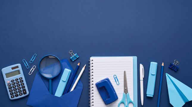 Top view flat lay of assorted blue school and office supplies including a notebook, calculator, envelope, and paper clips on a solid blue background.