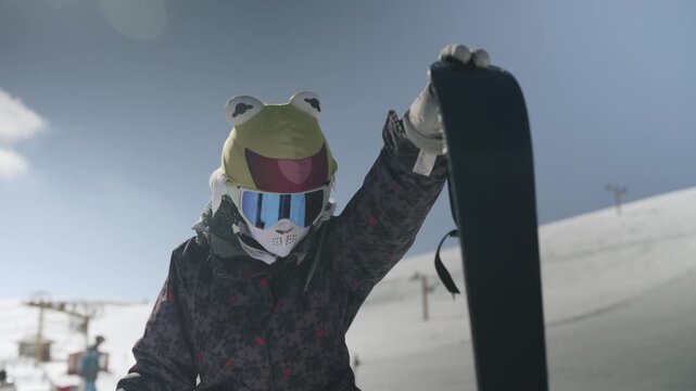A person wearing a playful frog hat and complete snow gear holds up a snowboard on a snowy mountain slope. This scene captures the spirit of winter sports and outdoor adventure.