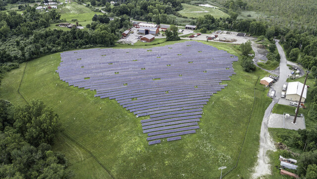 Aerial view of a large solar panel array installed in a grassy field, surrounded by rural barns and farm buildings in Patterson, New York, United States.