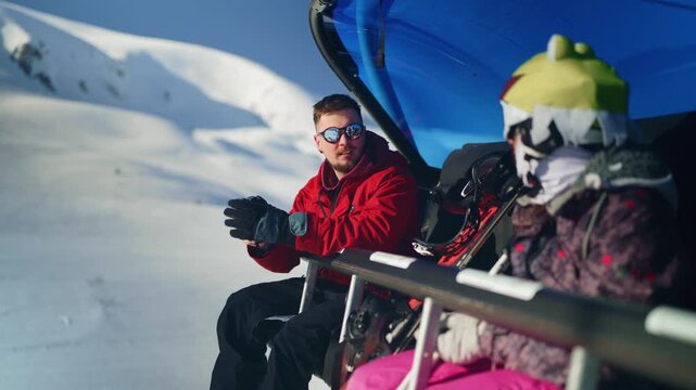 Two people, including a man in a red jacket, are seated on a ski lift ascending a snowy mountain. They are taking in the beautiful winter scenery during their ride up the mountain.