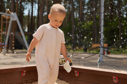 A young child explores a sand pit at a playground located in a park with tall trees around. The child holds a small bucket while playing in the sand